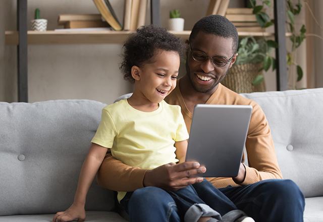 Excited loving african American dad hold tablet sit on couch with cute preschooler son watch cartoon together, smiling happy black dad and small boy kid relax on sofa at home play game on pad